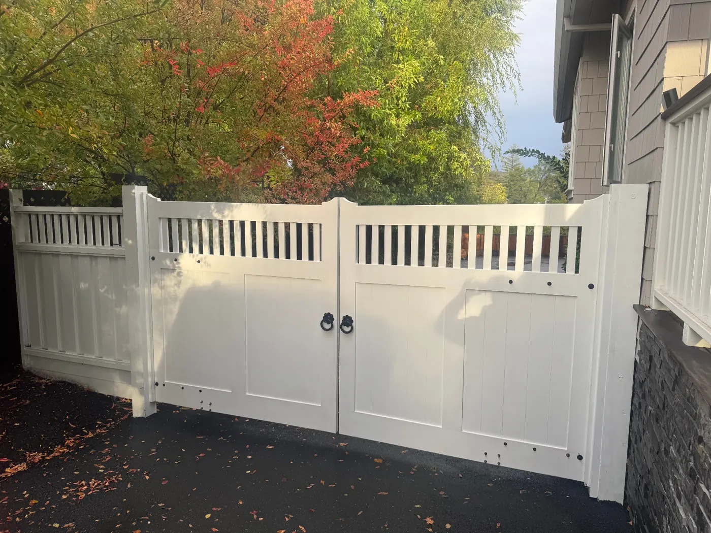 Automatic white-painted hardwood double driveway gate with spindle transom and iron ring pulls beside a Craftsman home