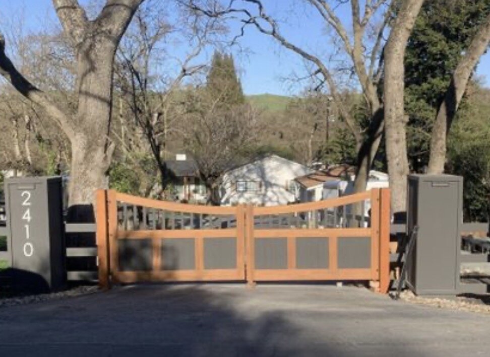 Arched wood and metal panel driveway gate at 2410 with automated operator pedestals under oak trees