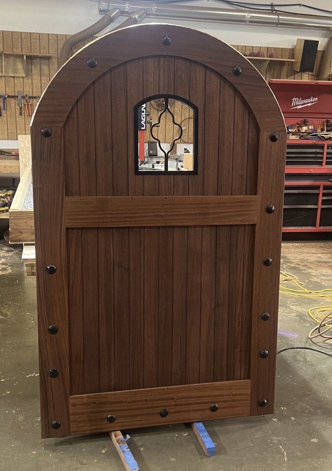 Arched mahogany gate with quatrefoil iron window and clavos detailing, shown in shop